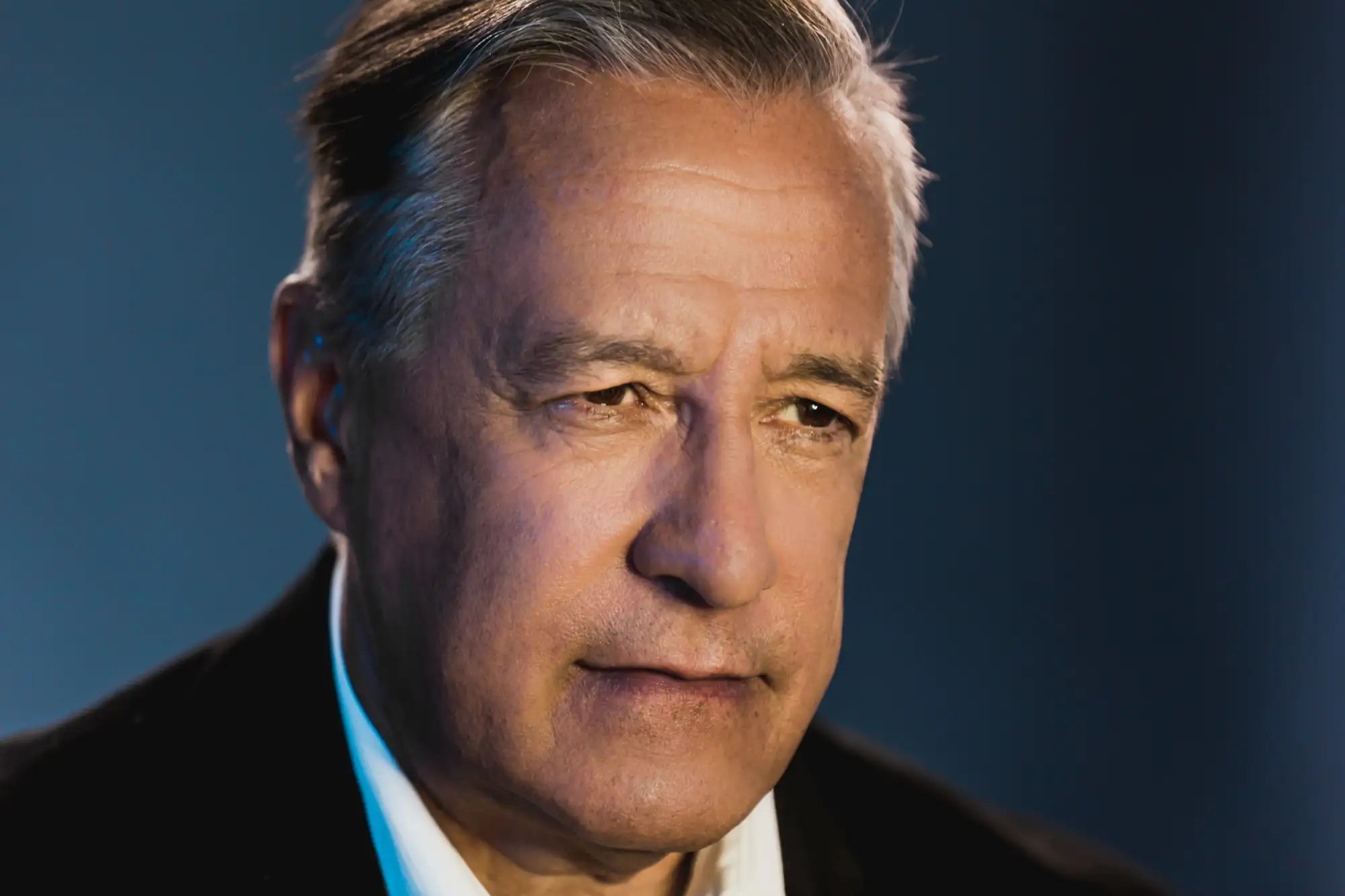 A close-up portrait of a man with gray hair, wearing a dark suit jacket over a white collared shirt.