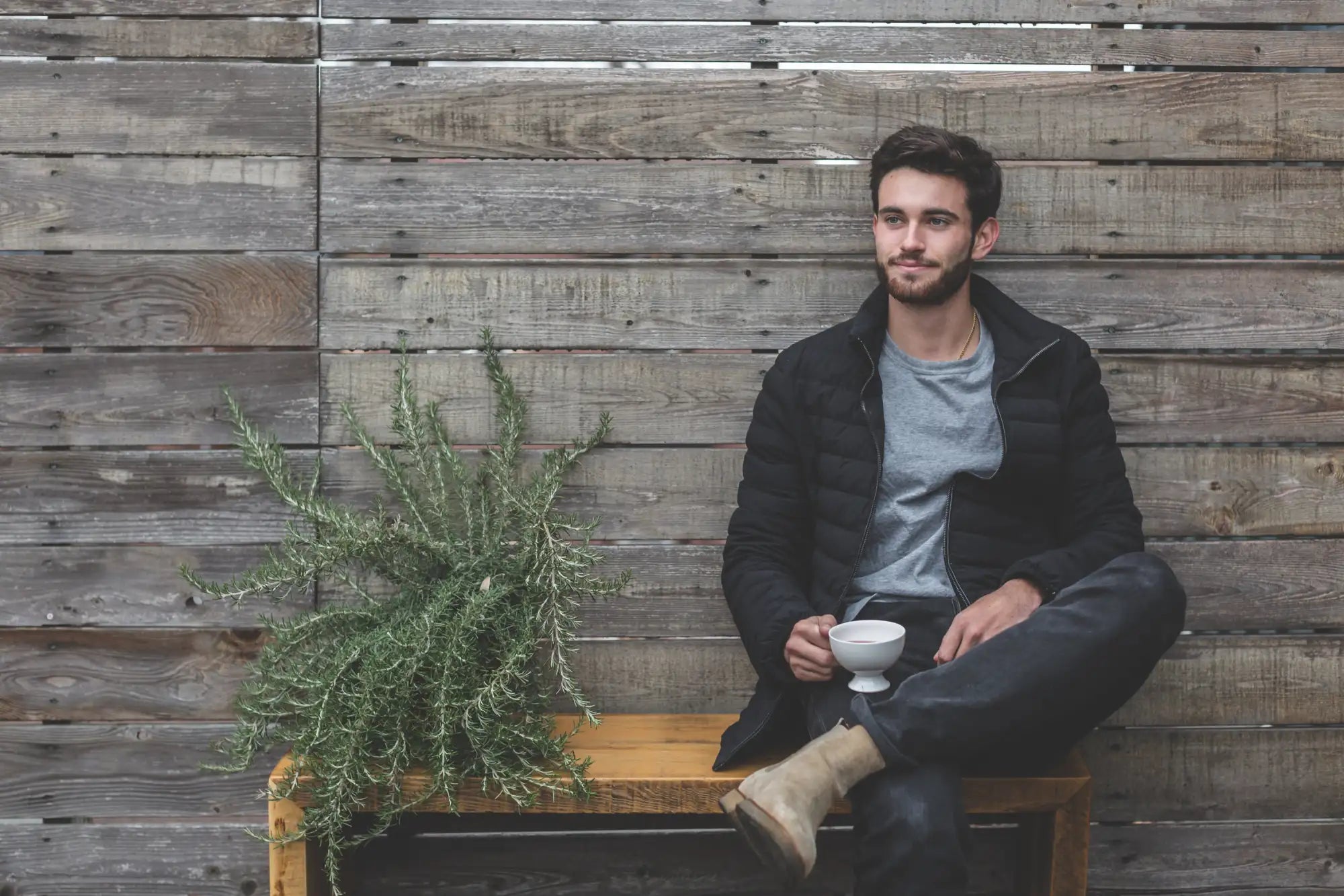 Man in black puffer jacket and grey t-shirt sitting on wooden bench holding white ceramic cup